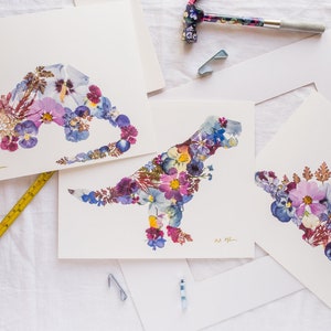 May include: Three framed prints of dinosaurs made from pressed flowers. The dinosaurs are in shades of purple, blue, and pink. The prints are signed "Art by Jill".