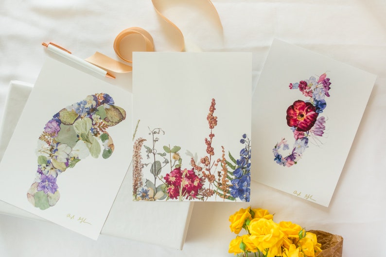 May include: Three framed prints with pressed flower art. The prints feature a manatee, a floral arrangement, and a seahorse. The prints are on white backgrounds and have a gold signature in the bottom right corner.