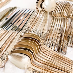 May include: A set of silver flatware, including knives, forks, and spoons, arranged on a white linen surface. The flatware has an ornate design with floral details.