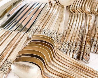 May include: A set of silver flatware, including knives, forks, and spoons, arranged on a white linen surface. The flatware has an ornate design with floral details.