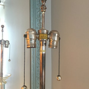 K&ouml;nnte beinhalten: Eine silberne Lampenbasis mit drei Lampenfassungen und schwarzen Kettenz&uuml;gen. Die Lampenbasis tr&auml;gt ein Etikett mit der Aufschrift "Manufactured Exclusively for The Mayfair Lamp Co.  Marked Lamp Co."