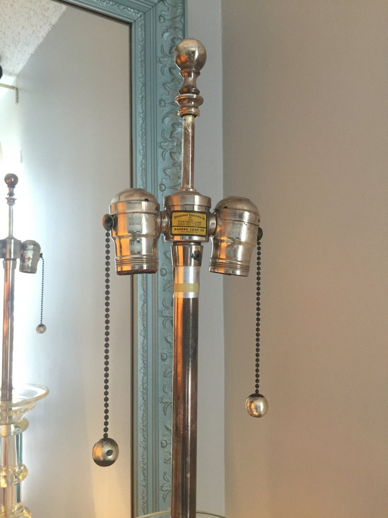 K&ouml;nnte beinhalten: Eine silberne Lampenbasis mit drei Lampenfassungen und schwarzen Kettenz&uuml;gen. Die Lampenbasis tr&auml;gt ein Etikett mit der Aufschrift "Manufactured Exclusively for The Mayfair Lamp Co.  Marked Lamp Co."