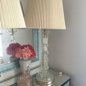 K&ouml;nnte beinhalten: Zwei Tischlampen mit gefalteten wei&szlig;en Lampenschirmen und klaren Glasf&uuml;&szlig;en. Die Lampen stehen auf einer verspiegelten Oberfl&auml;che mit einer Vase mit roten und rosa Blumen.