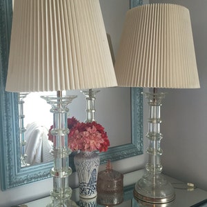K&ouml;nnte beinhalten: Zwei passende Glas-Tischlampen mit gefalteten wei&szlig;en Lampenschirmen. Die Lampen stehen auf einer Spiegelfl&auml;che mit einer Vase mit rosa Blumen und einem Dekorationsobjekt.