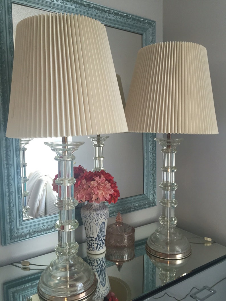 K&ouml;nnte beinhalten: Zwei passende Glas-Tischlampen mit gefalteten wei&szlig;en Lampenschirmen. Die Lampen stehen auf einer Spiegelfl&auml;che mit einer Vase mit rosa Blumen und einem Dekorationsobjekt.