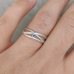 Könnte beinhalten: Ein silberner Ring mit einem blauen Edelstein in der Mitte. Der Ring hat ein einzigartiges Design mit mehreren sich verflechtenden Bändern.