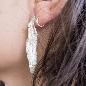 Puede incluir: Un par de pendientes de plata con un diseño texturizado y orgánico. Los pendientes son largos y cuelgan del lóbulo de la oreja.
