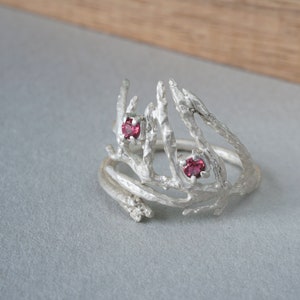 May include: A silver-colored ring with a unique, organic design. The ring features a textured, branch-like structure with two small, round, pink gemstones. The ring's design is reminiscent of natural forms.
