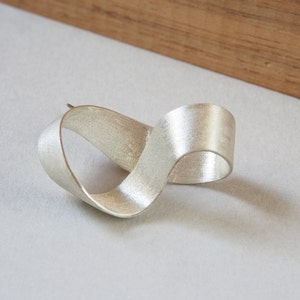 Puede incluir: Un broche de plata en forma de nudo infinito, hecho de una forma de cinta retorcida, se muestra sobre una superficie gris claro.