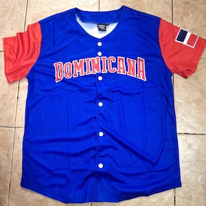 May include: A blue baseball jersey with red sleeves. The word "DOMINICANA" is across the chest in red letters. The Dominican Republic flag is on the right sleeve. The jersey has a v-neck and white buttons.