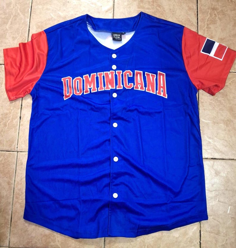 May include: A blue baseball jersey with red sleeves. The word "DOMINICANA" is across the chest in red letters. The Dominican Republic flag is on the right sleeve. The jersey has a v-neck and white buttons.