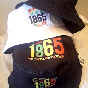 May include: Three bucket hats stacked on top of each other. The hats are black and white, with the text "1865" and "Juneteenth" in colorful fonts. The top hat has a graphic of three fists. The hats are on a white surface.