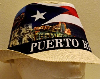 May include: A straw fedora hat with a band featuring the text "PUERTO RICO" in white letters. The hat's crown displays the Puerto Rican flag and a cityscape. The hat is beige and has a wide brim.