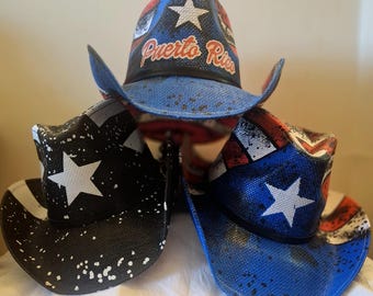 May include: Three cowboy hats featuring patriotic designs. The top hat is blue with a white star and the text Puerto Rico. The bottom left hat is black with white stars and paint splatters. The bottom right hat is blue with a white star and red stripes.