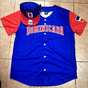 May include: A blue baseball jersey with red sleeves and the word "DOMINICANA" in red lettering. A matching blue and red baseball cap with the letters "RD" is placed on top of the jersey.