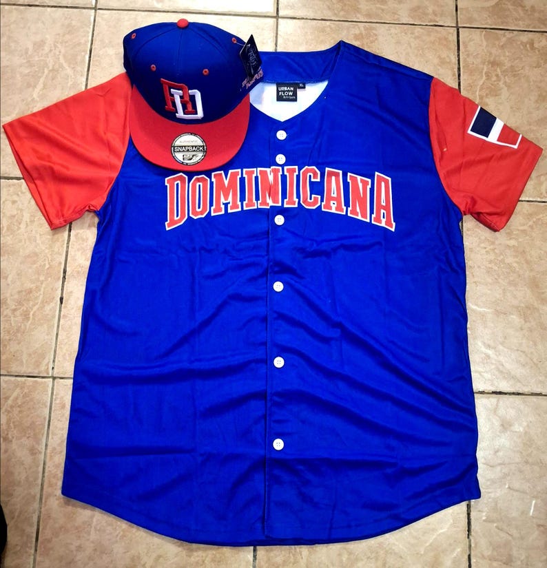 May include: A blue baseball jersey with red sleeves and the word "DOMINICANA" in red lettering. A matching blue and red baseball cap with the letters "RD" is placed on top of the jersey.