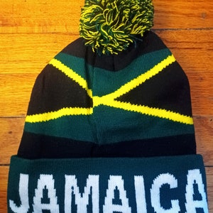 Peut inclure: Un bonnet en tricot noir, vert et jaune avec un pompon sur le dessus. Le bonnet porte l'inscription "JAMAICA" en blanc sur la bande inférieure.