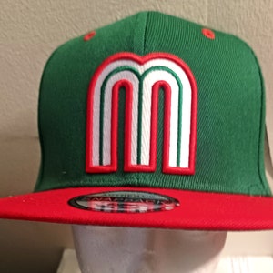 Könnte beinhalten: Grüne und rote Baseballkappe mit einem weißen und roten "M"-Logo. Die Kappe ist im Snapback-Stil.