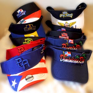 May include: A collection of nine blue, white, red, and black visor hats with embroidered logos and flags. The logos include "Dominican Republic", "Puerto Rico", "Virgin Islands", "Bahamas", and "KV Sports".