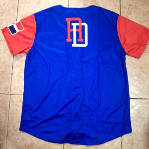 May include: A blue baseball jersey with red sleeves. The back features the letters "RD" in red with a white outline. The left sleeve has a Dominican Republic flag patch. The jersey is made of a shiny, athletic material.