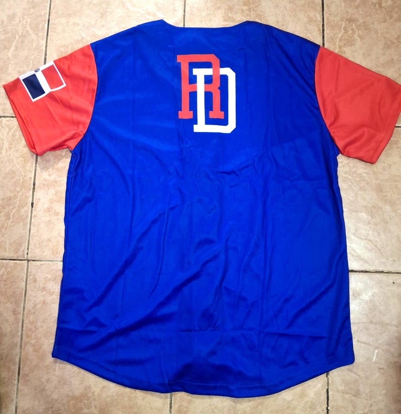 May include: A blue baseball jersey with red sleeves. The back features the letters "RD" in red with a white outline. The left sleeve has a Dominican Republic flag patch. The jersey is made of a shiny, athletic material.