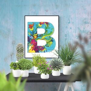May include: A framed print of the letter "B" with a colorful floral design on a blue background. The print is displayed on a table with several potted plants.