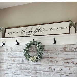 May include: A whitewashed wood wall with a shelf and hooks. The shelf has a sign that reads "Live Well Laugh Often Love Much". There is a wreath on the wall and a bench with a cushion that reads "Live Well Laugh Often Love Much".