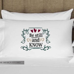 Pu&ograve; includere: Cuscino bianco con viti verdi e tre cuori rossi che circondano il testo "Be still and KNOW".