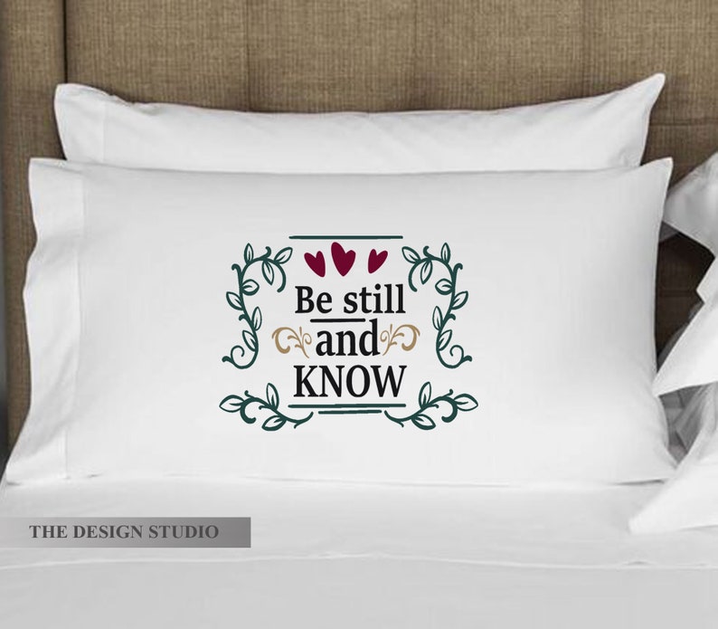 Pu&ograve; includere: Cuscino bianco con viti verdi e tre cuori rossi che circondano il testo "Be still and KNOW".