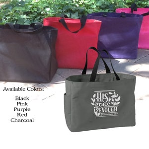 May include: A collection of tote bags in various colors: black, pink, purple, red, and charcoal. The front-most bag is charcoal with a white graphic that reads "His grace is enough" and a Bible verse reference. Each bag has black handles.