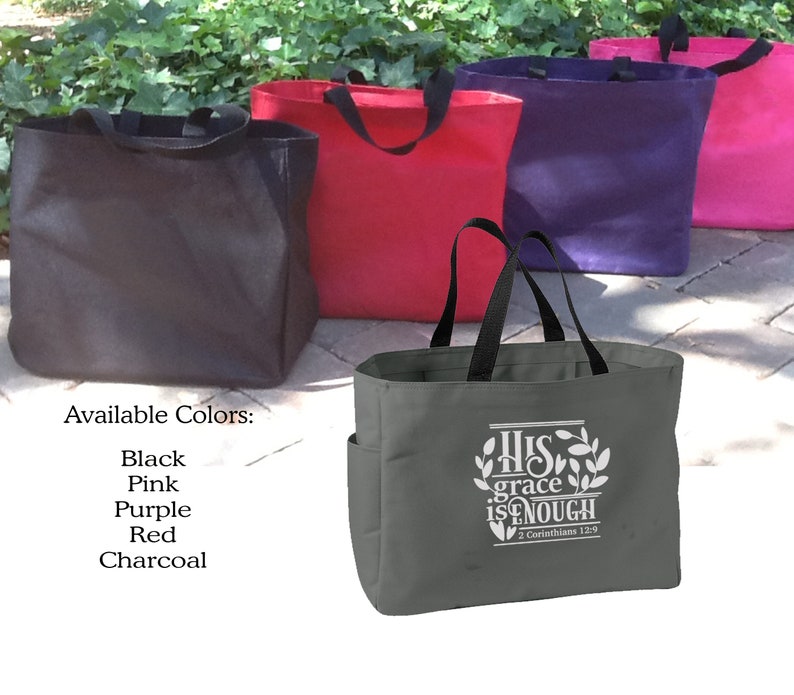 May include: A collection of tote bags in various colors: black, pink, purple, red, and charcoal. The front-most bag is charcoal with a white graphic that reads "His grace is enough" and a Bible verse reference. Each bag has black handles.