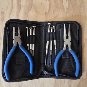 May include: A black zippered tool kit containing a set of pliers with blue handles, a set of needle-nose pliers with blue handles, and several small screwdrivers. The tools are arranged against a light wood grain background.