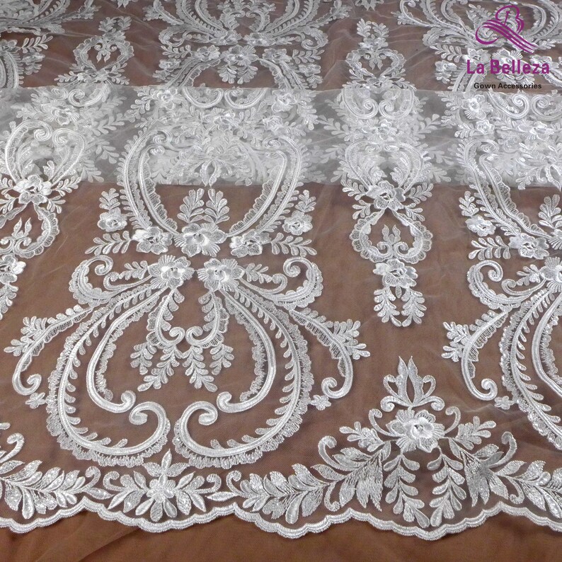 K&ouml;nnte beinhalten: Wei&szlig;er Spitzenstoff mit einem zarten Blumenmuster. Die Spitze ist mit kleinen wei&szlig;en Perlen und Pailletten bestickt. Der Stoff ist durchsichtig und hat eine weiche, flie&szlig;ende Textur.