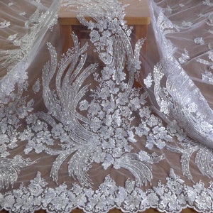 May include: White tulle fabric with silver glitter and floral embroidery. The fabric has a delicate, feminine look and would be perfect for a wedding dress or other special occasion garment.