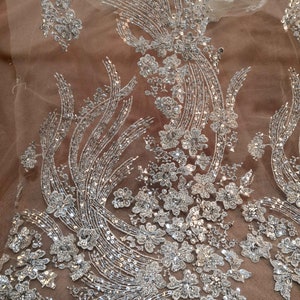 May include: Silver sequined fabric with a floral design on a beige mesh background. The fabric is perfect for adding a touch of sparkle to any project.