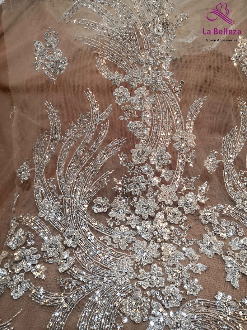 May include: Silver sequined fabric with a floral design on a beige mesh background. The fabric is perfect for adding a touch of sparkle to any project.