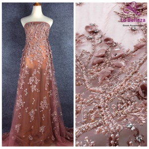 May include: A close-up of a delicate, blush-colored lace fabric with intricate floral patterns and pearl embellishments. The fabric is sheer and has a soft, flowing texture. The text "La Belleza Gown Accessories" is visible in the upper right corner of the image.