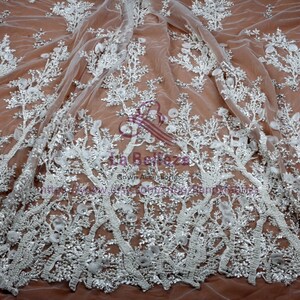 May include: White floral lace fabric with beaded embellishments. The lace features a delicate floral pattern with 3D flowers and intricate beading. The fabric is sheer and lightweight, making it ideal for special occasion garments.