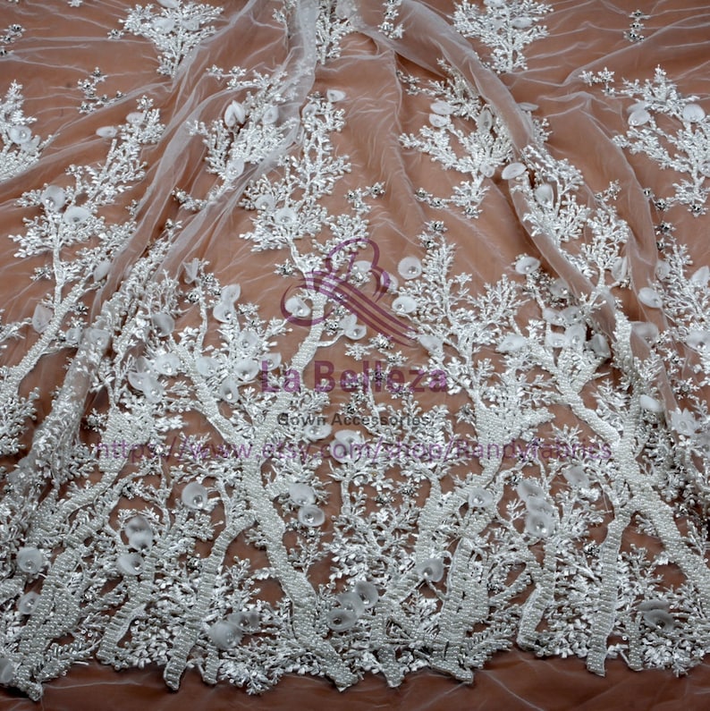 May include: White floral lace fabric with beaded embellishments. The lace features a delicate floral pattern with 3D flowers and intricate beading. The fabric is sheer and lightweight, making it ideal for special occasion garments.