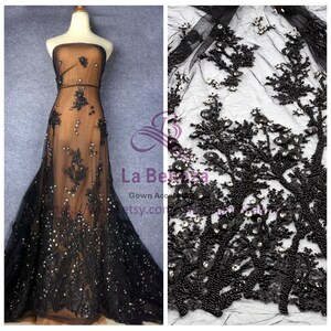 May include: Black beaded lace fabric with a floral design. The lace is on a sheer mesh background. The fabric is shown on a dress form and as a close-up.