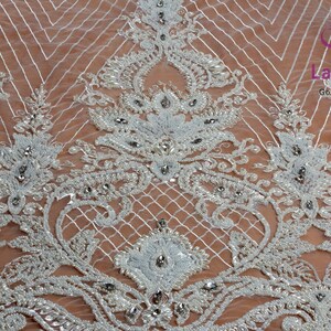 May include: White beaded lace fabric with a delicate floral pattern and small rhinestones. The fabric has a sheer, lightweight texture and is perfect for adding a touch of elegance to any project.