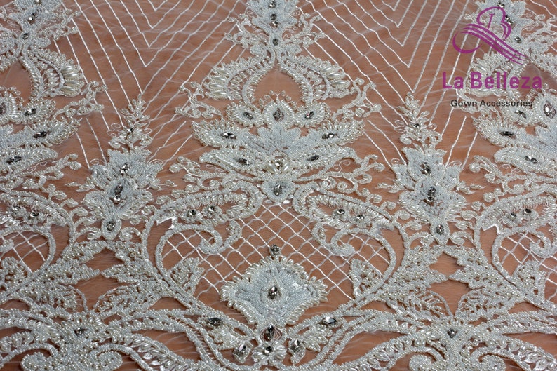 May include: White beaded lace fabric with a delicate floral pattern and small rhinestones. The fabric has a sheer, lightweight texture and is perfect for adding a touch of elegance to any project.