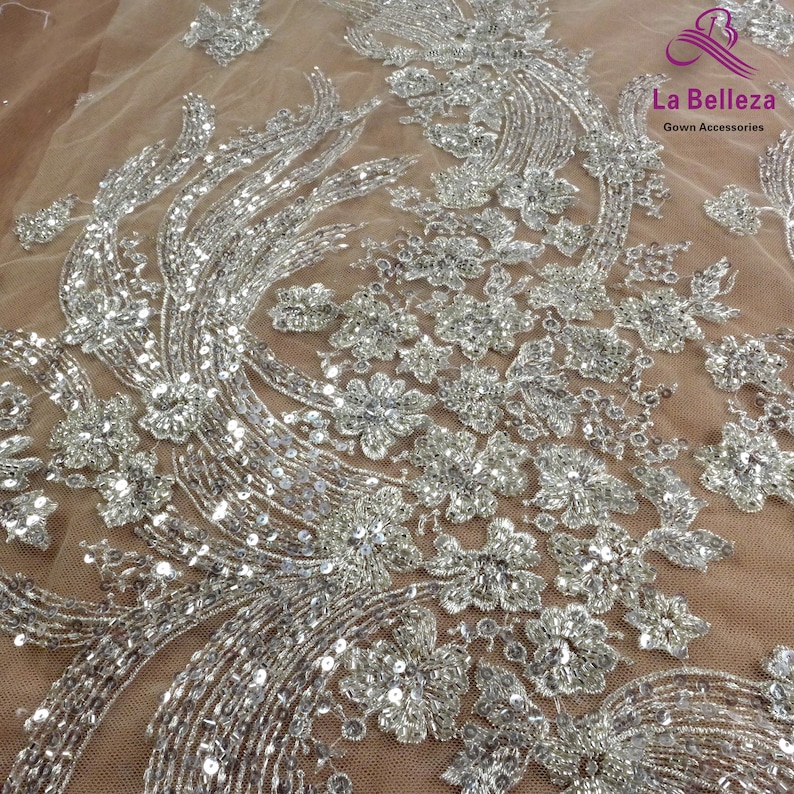May include: Silver sequined fabric with a floral design on a sheer beige mesh background. La Belleza Gown Accessories is printed in the top right corner.