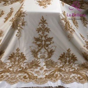 Op de afbeelding: Gouden kralen kant stof met een bloemenpatroon. De stof is doorschijnend en heeft een delicate textuur. La Belle Gown Accessories is op de stof gedrukt.
