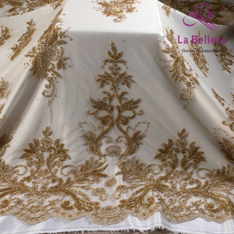 Op de afbeelding: Gouden kralen kant stof met een bloemenpatroon. De stof is doorschijnend en heeft een delicate textuur. La Belle Gown Accessories is op de stof gedrukt.