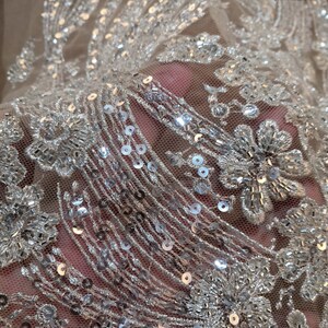 May include: Close-up of a sheer fabric with silver sequins and beading. The fabric has a floral pattern and is perfect for adding a touch of sparkle to any project.