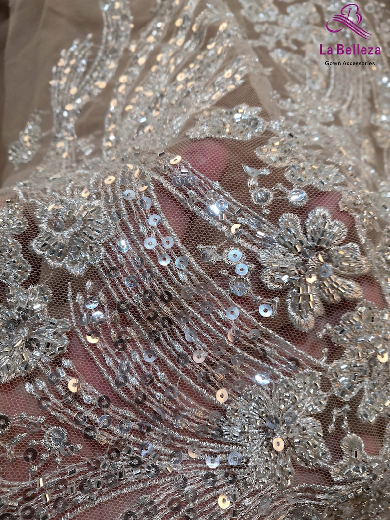 May include: Close-up of a sheer fabric with silver sequins and beading. The fabric has a floral pattern and is perfect for adding a touch of sparkle to any project.