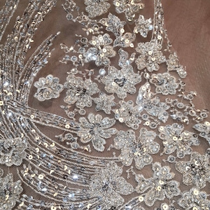 May include: Close-up of a silver sequined fabric with a floral design. The fabric is sheer and has a delicate, feminine look.
