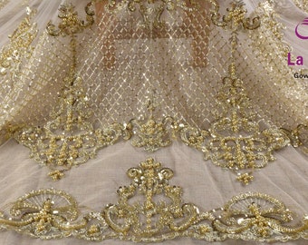 May include: Gold beaded lace fabric with a delicate pattern. The fabric is sheer and has a sparkly finish. La Belleza Gown Accessories is printed on the fabric.