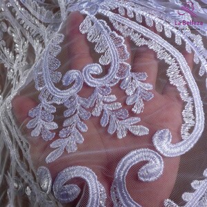 K&ouml;nnte beinhalten: Wei&szlig;er Spitzenstoff mit einem zarten Blumenmuster. Die Spitze ist mit wei&szlig;em Garn bestickt und hat eine weiche, durchscheinende Textur.
